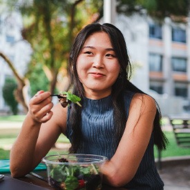 Woman with black hair in blue shirt eating salad outside at park table