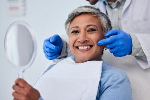 Woman with short gray hair in dentist's chair smiling at the camera holding a mirror